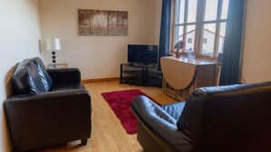 Un salon avec deux canapés en cuir noir, une table basse en bois, un tapis rouge, un téléviseur à écran plat, un tableau accroché au mur et une fenêtre avec des rideaux.
