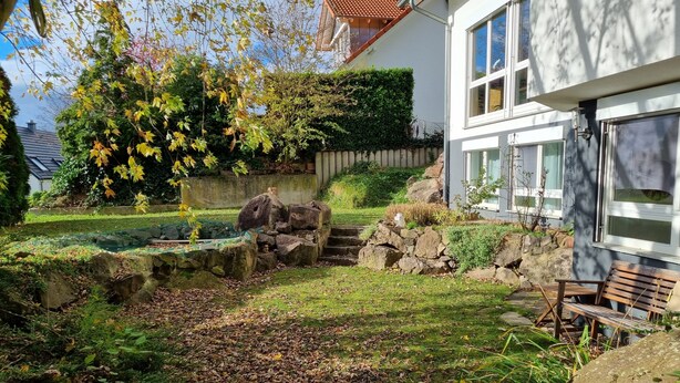 A garden with a stone wall, a wooden bench, and a house with a white facade and large windows.