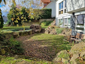 En trädgård med en stenmur, en träbänk och ett hus med en vit fasad och stora fönster.