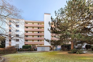 Un immeuble résidentiel à plusieurs étages, doté d’une tour centrale blanche, entouré d’une zone verdoyante et d’arbres.