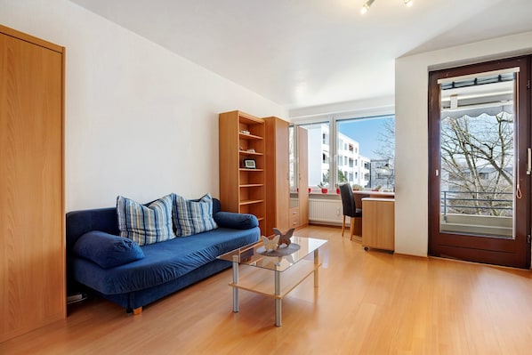 Un salon moderne avec un canapé bleu, une table basse en verre, une bibliothèque en bois et une vue dégagée sur l’extérieur grâce à une grande fenêtre.