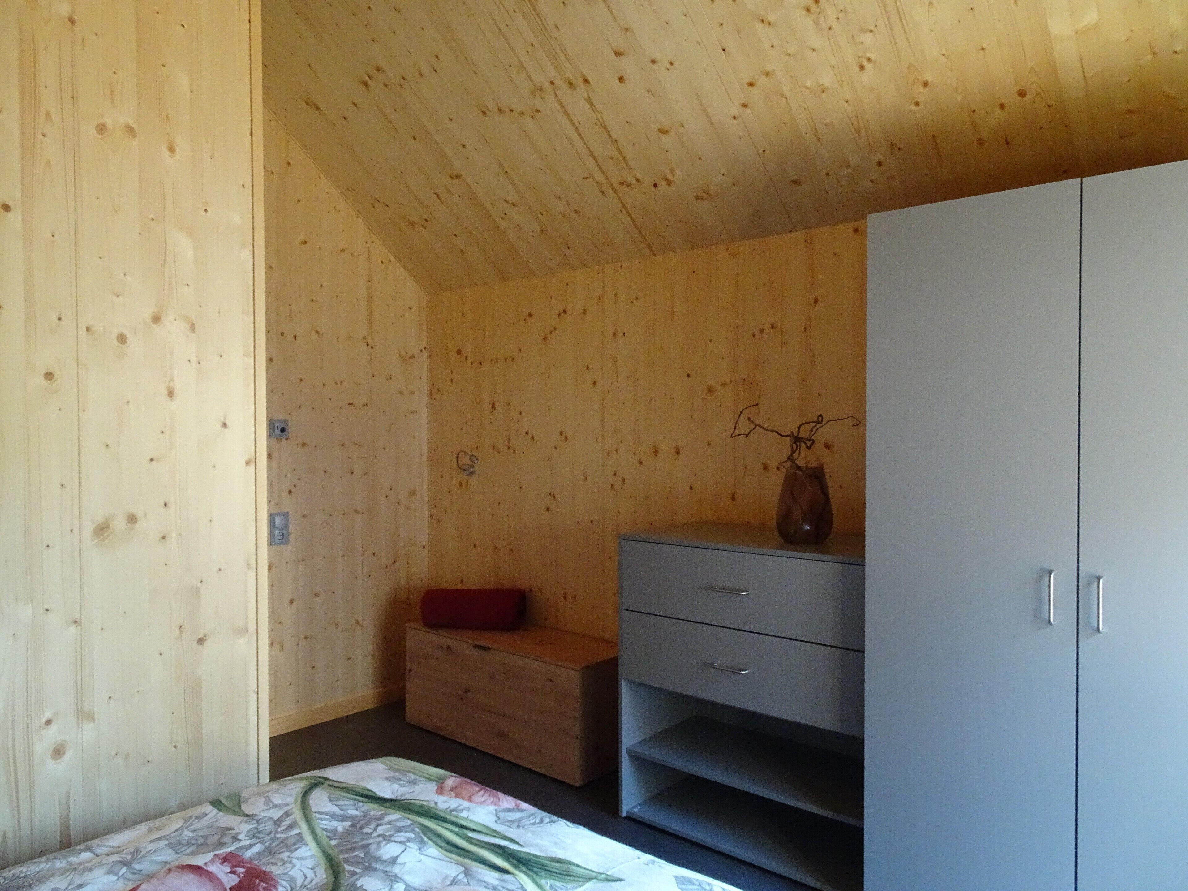 Une pièce aux murs en bois, avec un lit recouvert d’une literie à motifs floraux, une commode et une armoire.