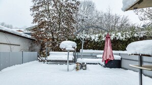 Un patio trasero cubierto de nieve, con una cerca metálica, un árbol y una sombrilla.