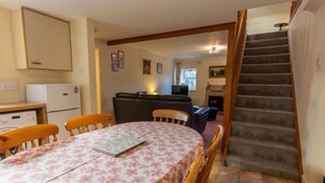 A dining area with a round table covered in a floral tablecloth, wooden chairs, and a kitchen area with a refrigerator and stove. - Old Lodge by Interhome (Inverness)