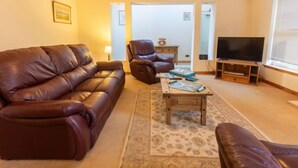 A living room with a brown leather sofa, a brown leather armchair, a wooden coffee table with board games, a television on a wooden stand, and a patterned rug. - Lodge Road by Interhome (Inverness)