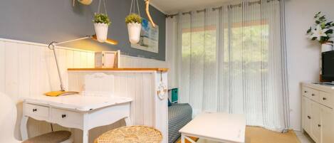 A compact room with a white desk, a chair, a small table, a wicker basket, and a bed with a white headboard.
