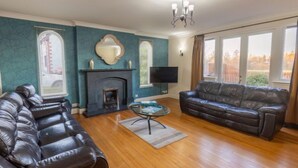 A living room with a fireplace, leather sofas, a flat-screen TV, and large windows. - Evergreen House by Interhome (Inverness)