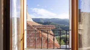 Une vue depuis une fenêtre donnant sur un balcon, avec en contrebas un bâtiment dont le toit est recouvert de tuiles rouges et, au loin, un paysage montagneux.
