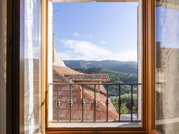 A view from a window with a balcony, overlooking a building with a red-tiled roof and a mountainous landscape in the background.