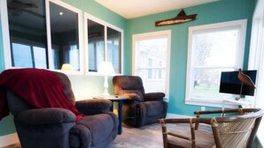 Une pièce aux murs de couleur turquoise, avec deux fauteuils sombres, une chaise en bois, une petite table avec une lampe et un téléviseur posé sur un meuble.