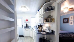 Un couloir étroit, avec un coin cuisine sur la droite, comprenant des meubles blancs, un four à micro-ondes et divers ustensiles de cuisine. Une porte se trouve au fond du couloir et donne accès à une autre pièce.