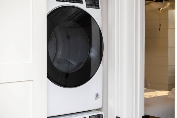 A front-loading washing machine in a room with a bed and a hanging light fixture.