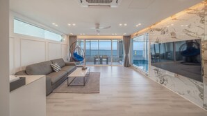 A modern living room with a sofa, coffee table, and hanging chair, large windows with a view of the sea, and a marble wall. - Pohang Etoile Poolvilla (Pohang)
