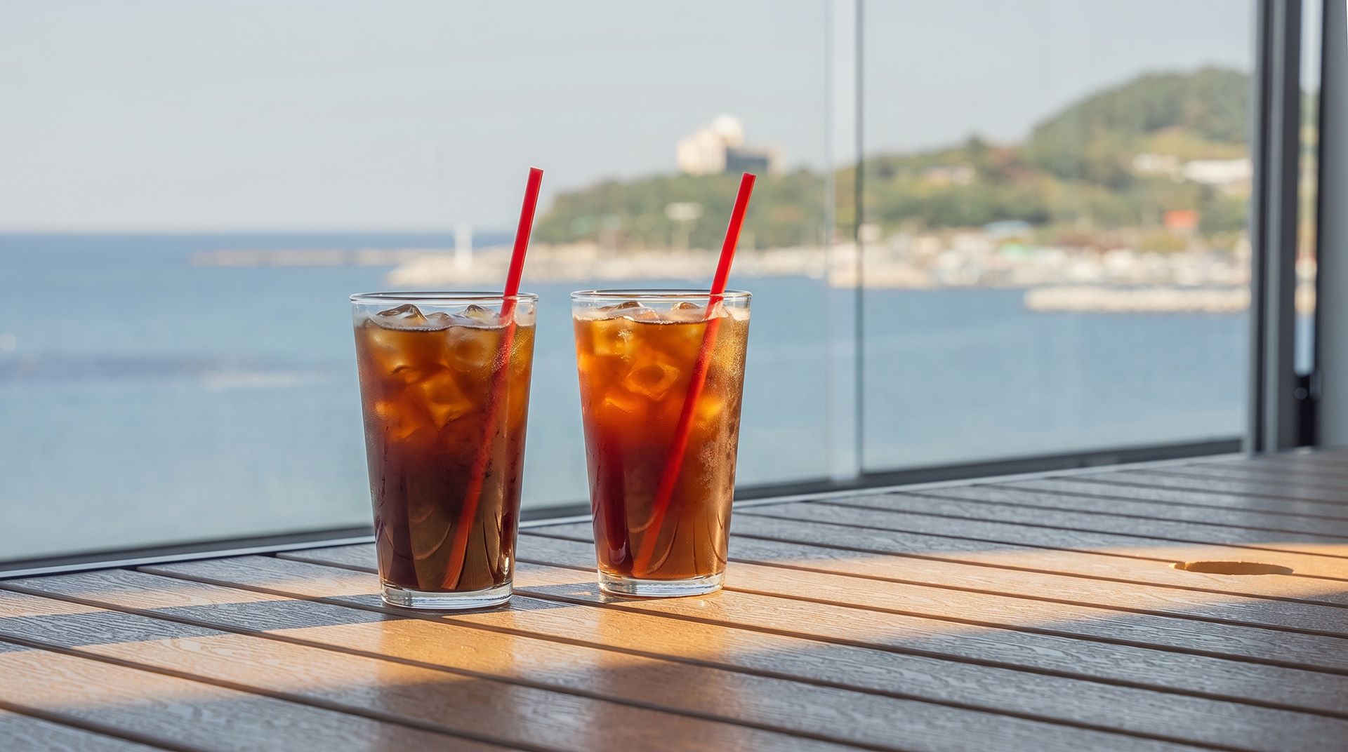 Two glasses of iced tea on a wooden table with a view of the sea and a distant hill.