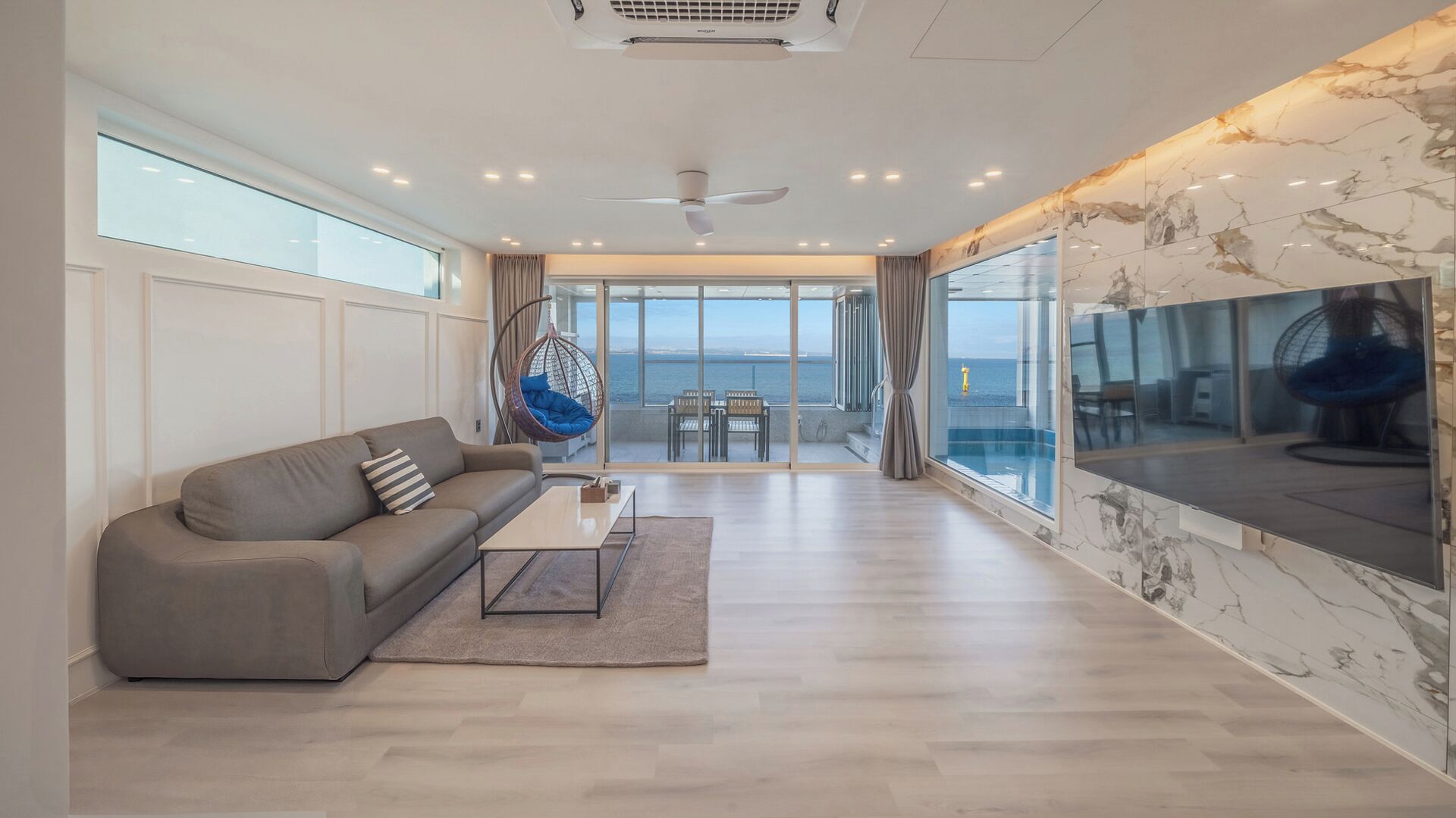 A modern living room with a sofa, coffee table, and hanging chair, large windows with a view of the sea, and a ceiling fan.