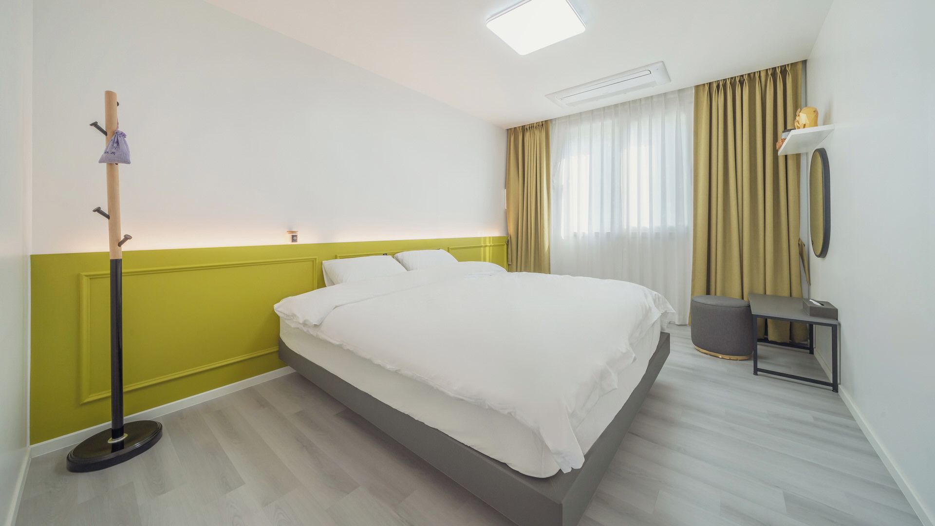 Ein Hotelzimmer mit Bett, Nachttisch, Lampe, Kleiderhaken und einem Fenster mit Vorhängen.