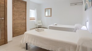 Een hotelkamer met een houten deur, een witte stoel, twee bedden, een raam met gordijn, een spiegel aan de muur en een schilderij.