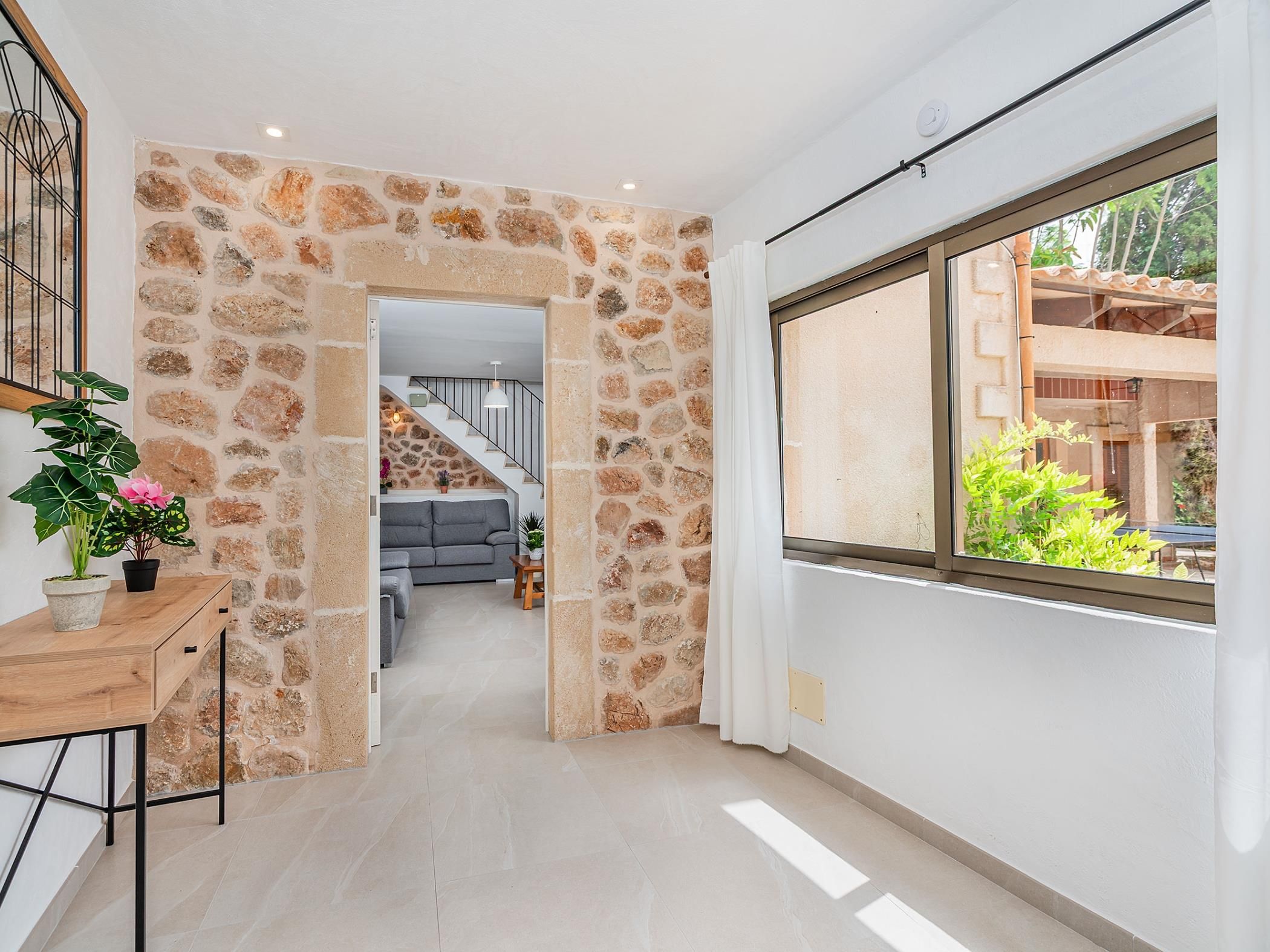 A hallway with a stone wall, a wooden desk with a plant, and a view of a living room with a sofa and a staircase.