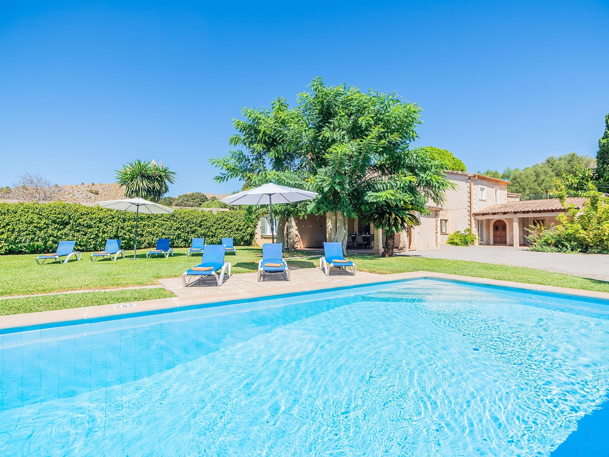 A swimming pool with lounge chairs and umbrellas, surrounded by greenery and a building.