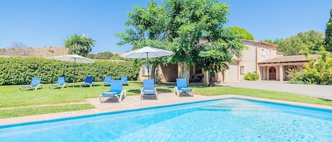 A swimming pool with lounge chairs and umbrellas, surrounded by greenery and a building.