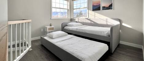 A bedroom with a bunk bed, a single bed, a nightstand, a crib, and two windows with a view of mountains.