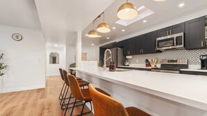 Una cocina moderna con encimera blanca, armarios oscuros y un microondas de acero inoxidable. Hay tres lámparas colgantes sobre la encimera y un reloj en la pared.