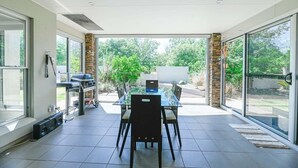 Un'ampia veranda con tavolo e sedie in vetro, una griglia per barbecue e vista sul verde.