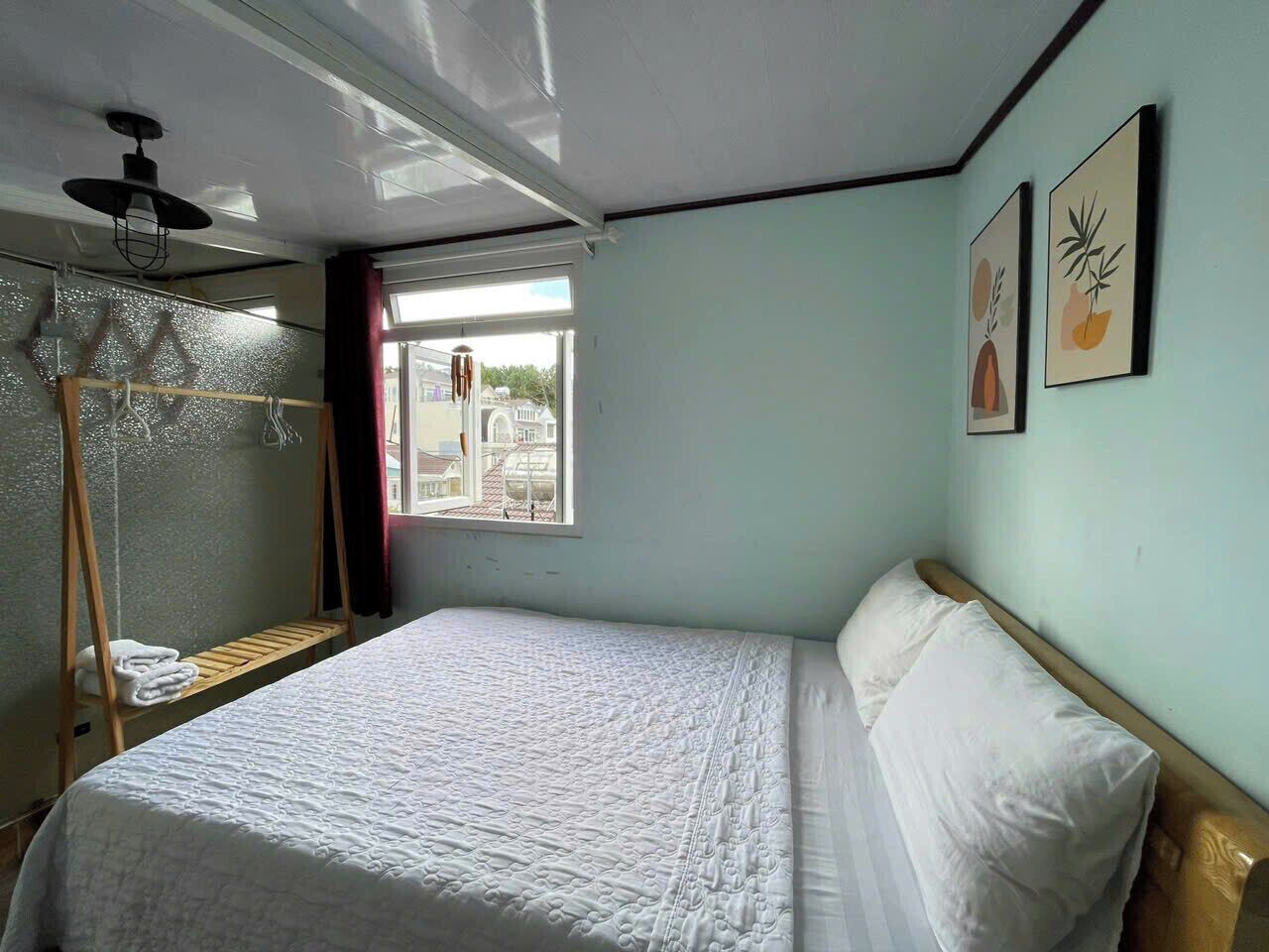 A bedroom with a bed, a wooden shelf, a hanging light fixture, and a window with a view of buildings outside.