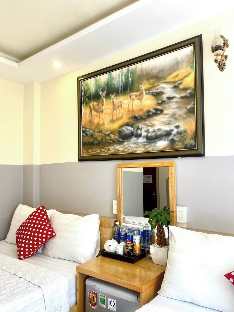 A hotel room with a bed, a bedside table with a plant, a mirror, and a painting of deer by a stream.