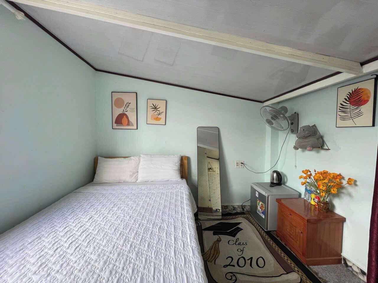 A bedroom with a bed, a dresser, a mirror, and a small desk. There are wall decorations and a fan.