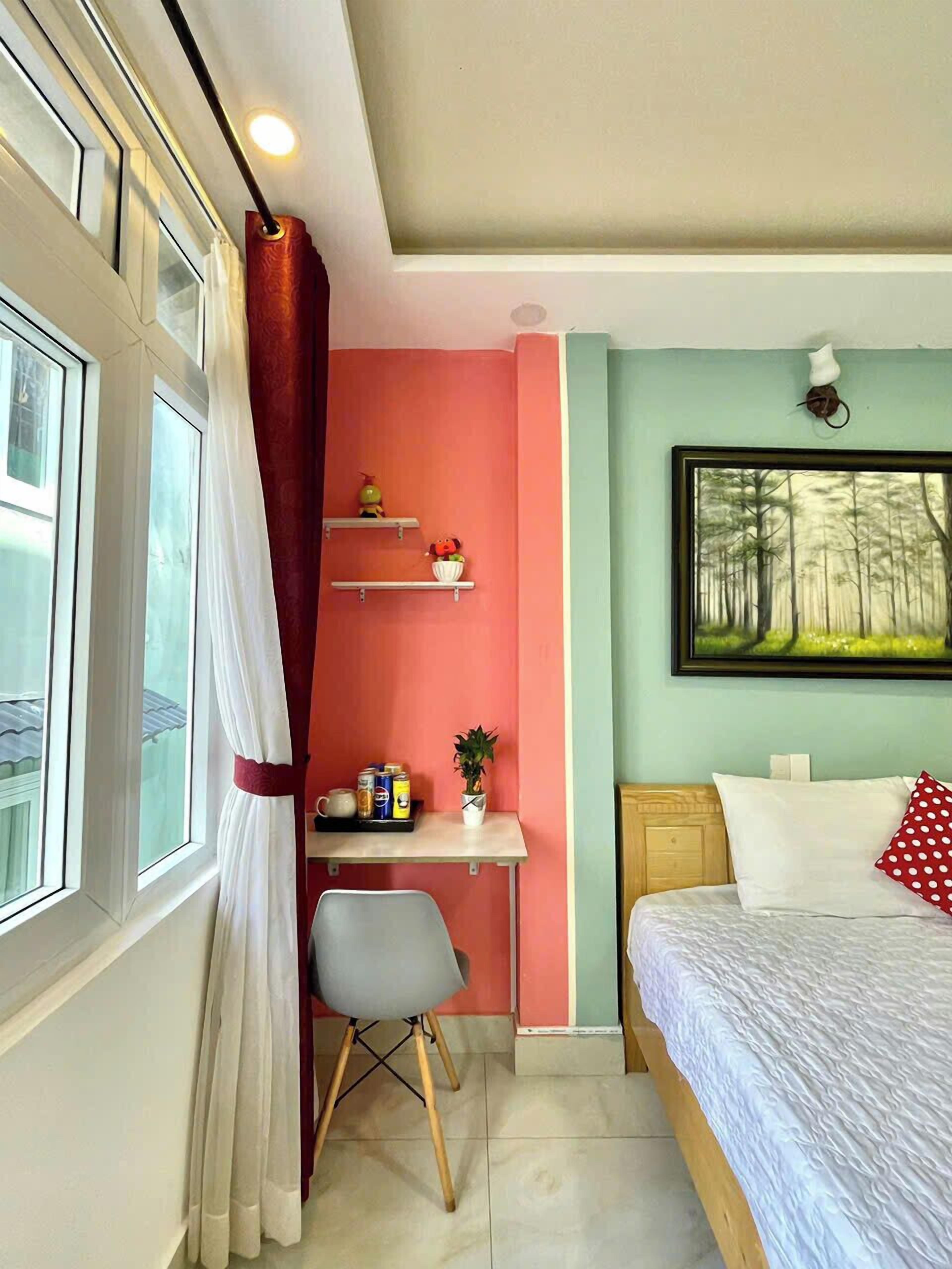 A bedroom with a bed, a desk with a chair, a shelf with decorative items, a window with curtains, and a painting on the wall.