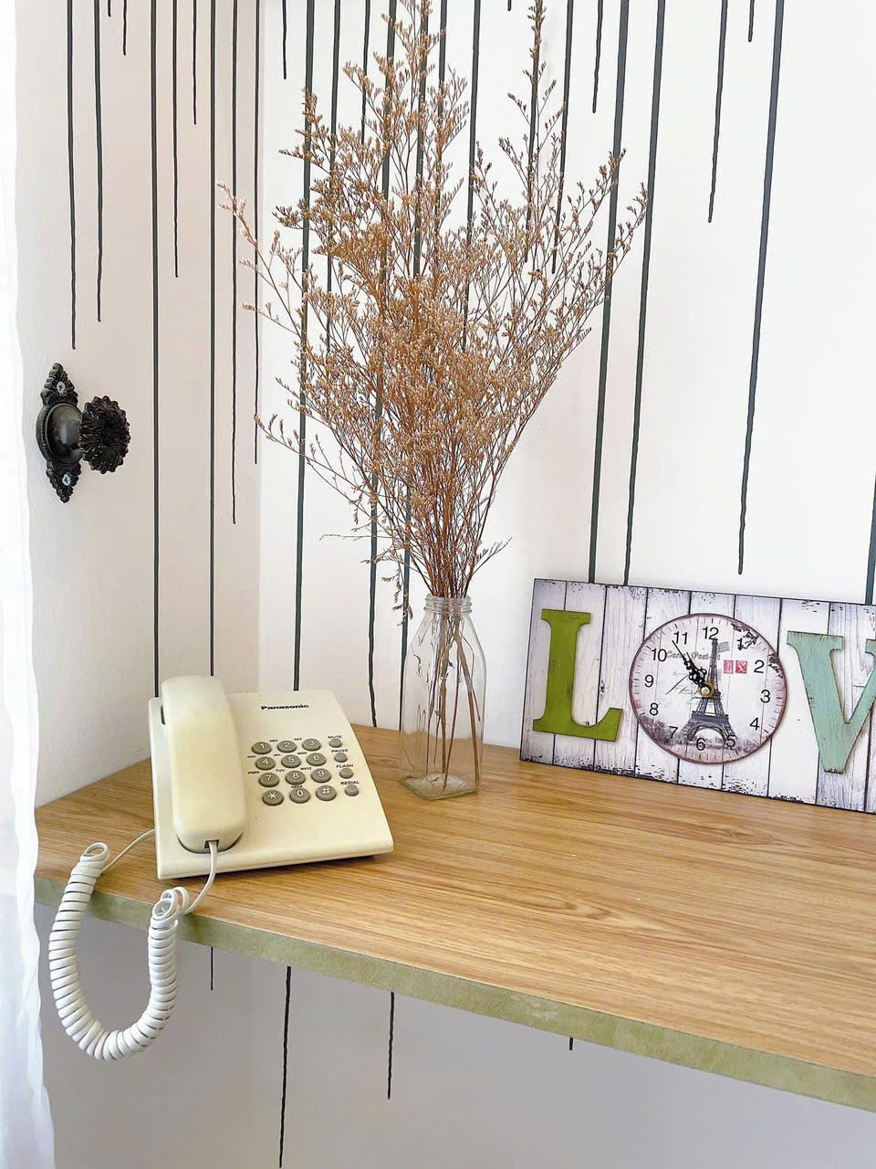 Una reception in legno con un telefono a disco bianco, un vaso con rami secchi e una sveglia a muro con le lettere 'L' e 'V'.
