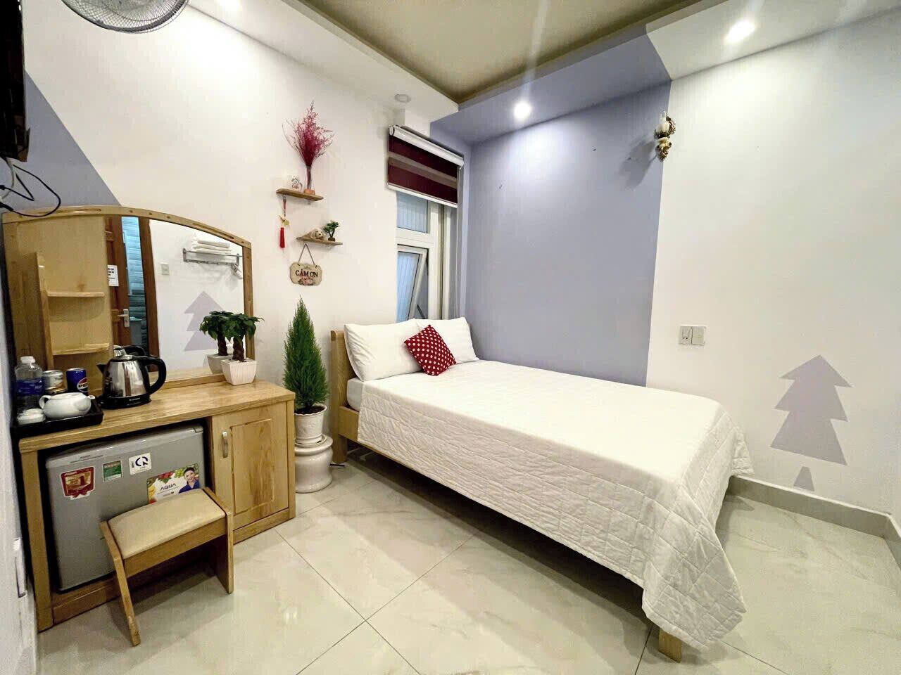 Quarto compacto com cama, uma pequena mesa com chaleira, geladeira e planta em vaso.
