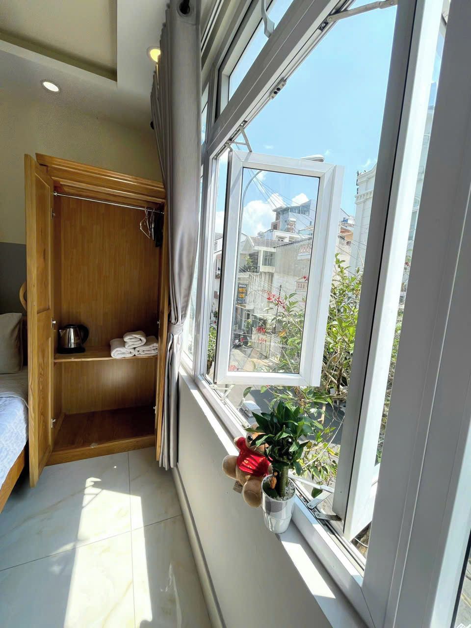 Una habitación con un armario de madera, una cama, un ventanal con vistas a edificios y zonas verdes, y una pequeña planta en maceta en el alféizar.