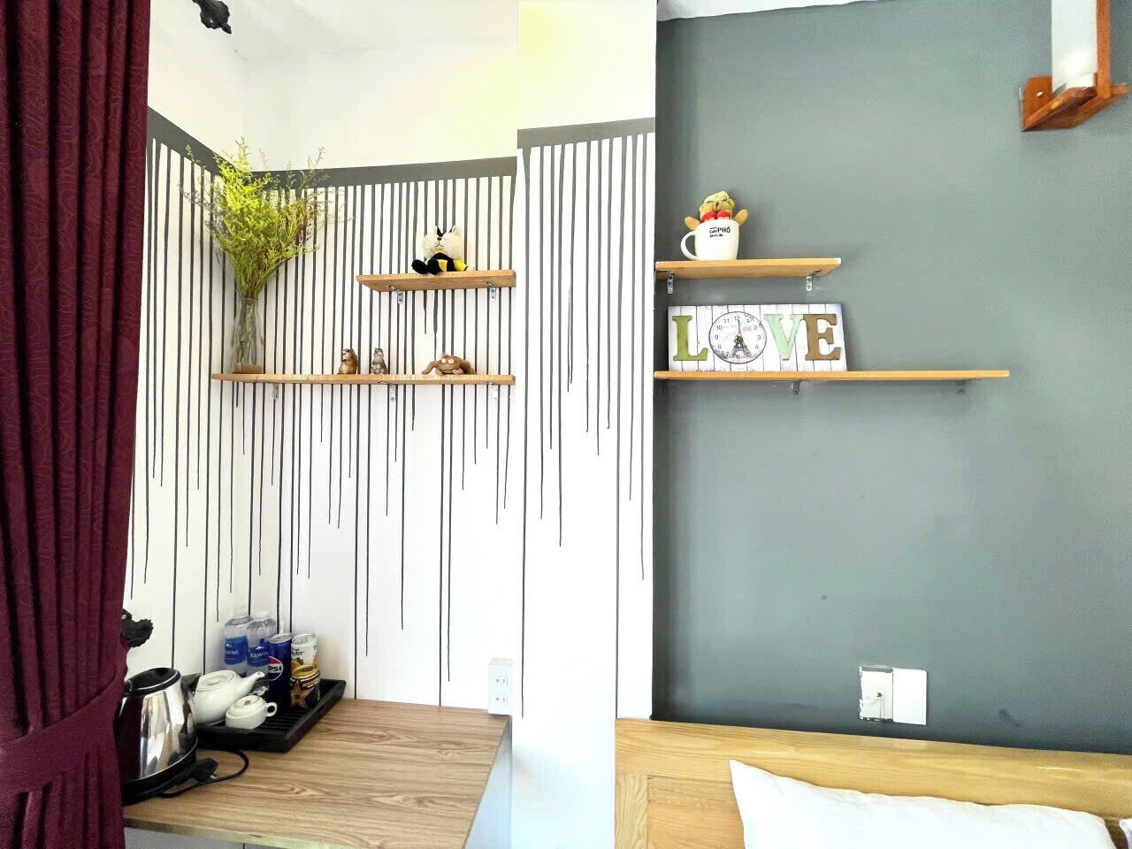 A room with a wooden bed, a small table with a kettle and teapot, a shelf with decorative items, and a wall with a 'LOVE' sign.