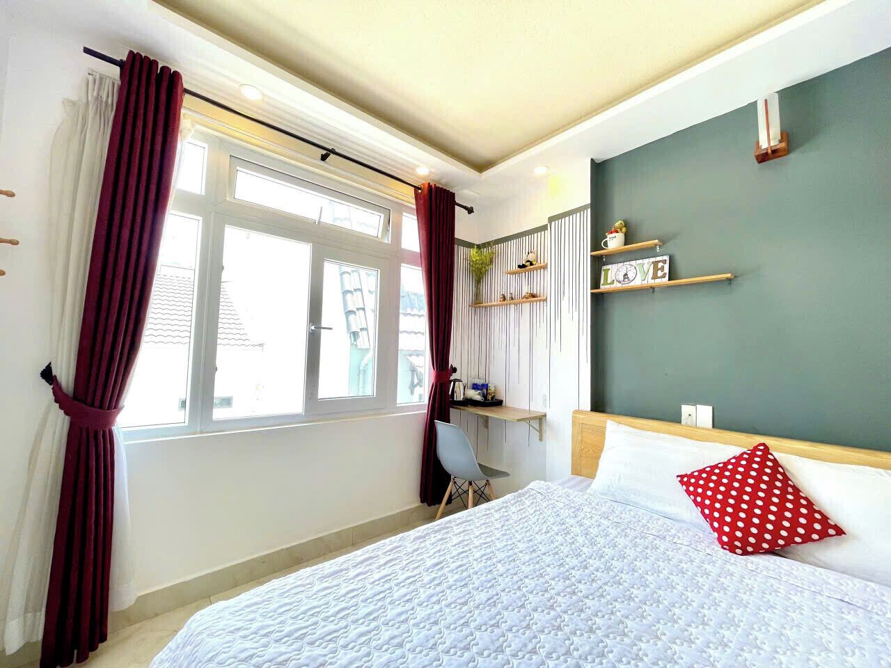 A bedroom with a bed, a desk, and a chair. There is a window with curtains, a shelf with items, and a wall with a sign.