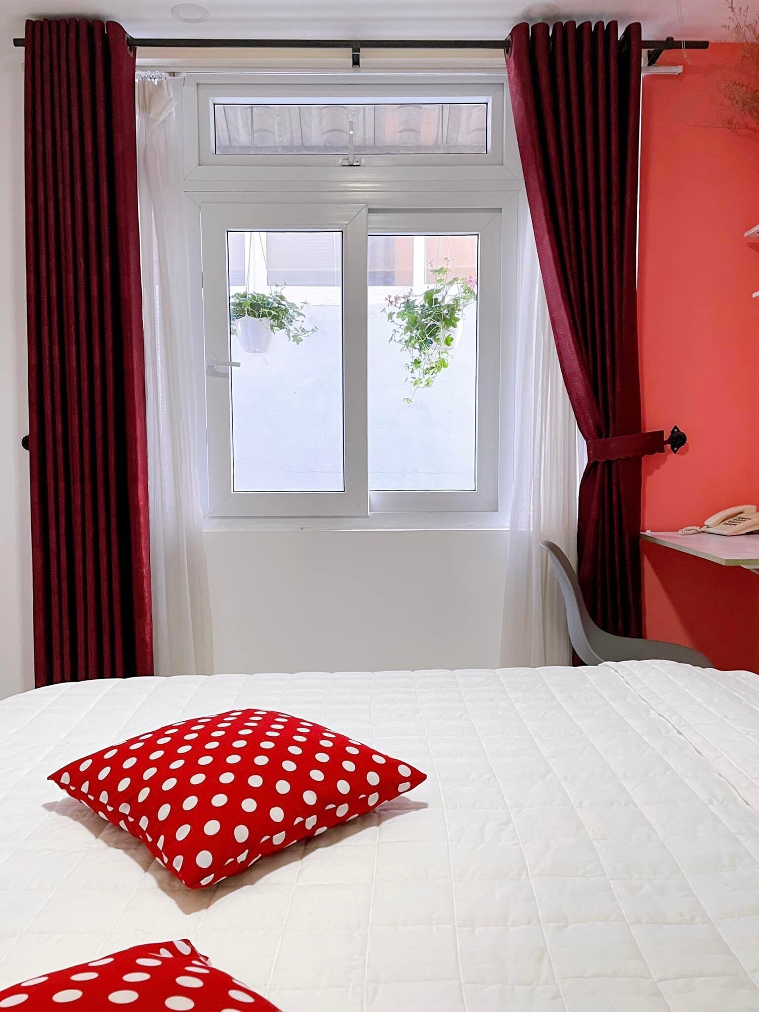 A hotel room with a bed, red curtains, a window with a view of greenery, and a telephone on a desk.