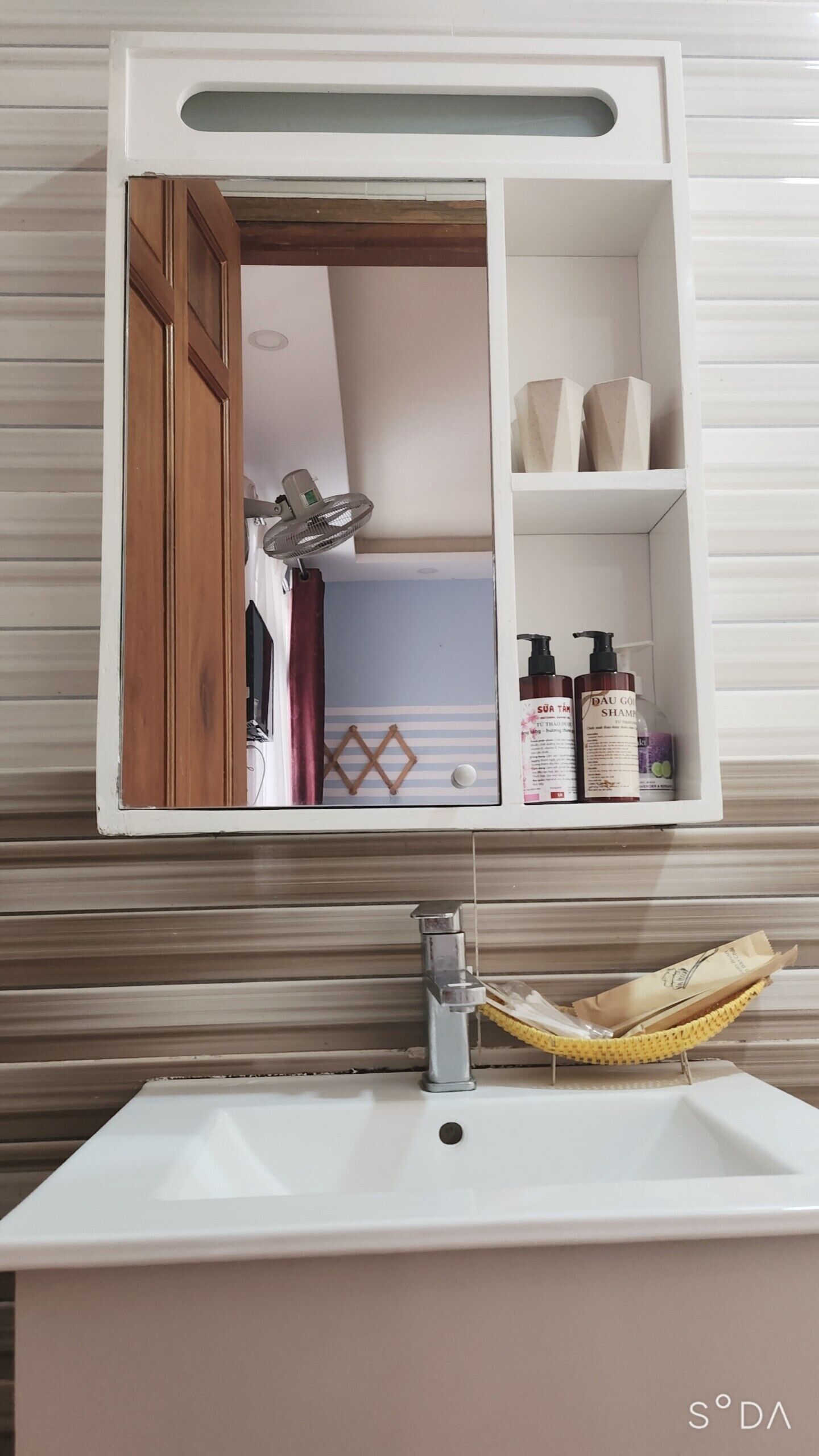 Un baño con lavamanos blanco, un espejo y un estante con artículos de aseo.