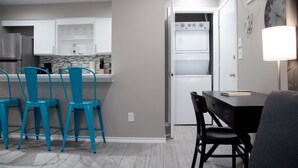 A modern kitchen with white cabinets, a built-in oven, and a dining area with a black table and blue chairs. - Montrose Chelsea Studio! Medical Center 10 (Houston)