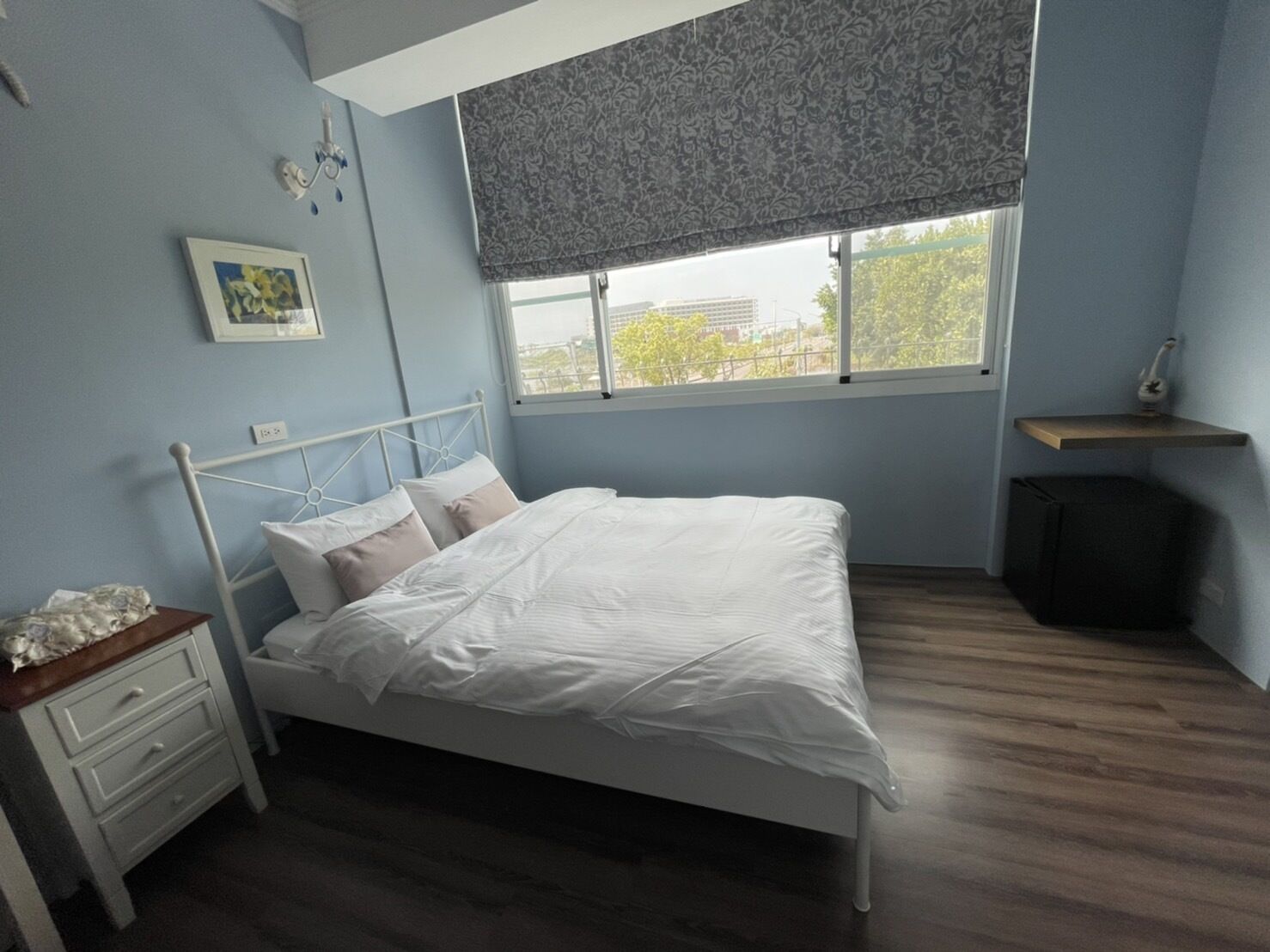 A bedroom with a bed, a nightstand, a dresser, a window with a view, and a bedside table.