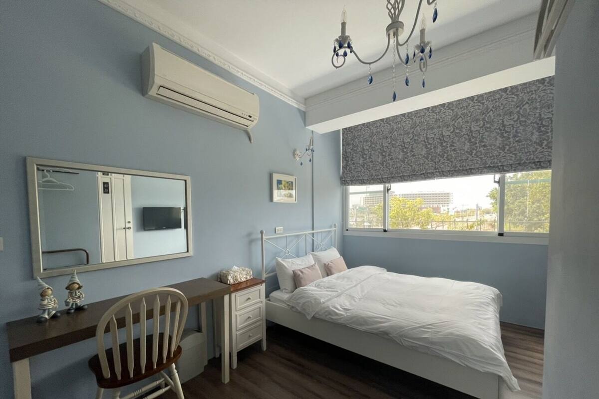 A bedroom with a bed, a desk, a chair, a dresser, a mirror, and a window with a view.