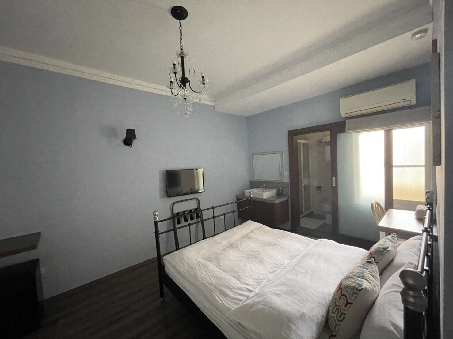 A bedroom with a bed, a bedside table, a mirror, a TV, and an air conditioning unit.