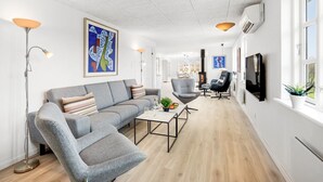 Ein modernes Wohnzimmer mit einem grauen Sofa, einem Couchtisch und einem Fernseher. Zwei Sessel, eine Stehlampe und Kunstwerke an den Wänden ergänzen das Bild.