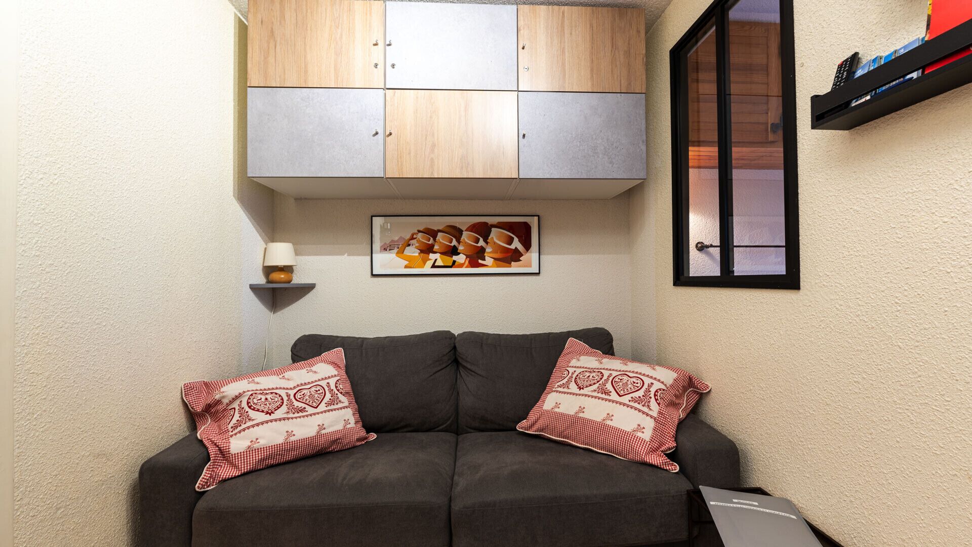 Un salon avec un canapé gris, deux coussins décoratifs à motifs, un tableau encadré, une petite étagère avec une lampe et une fenêtre avec des stores.