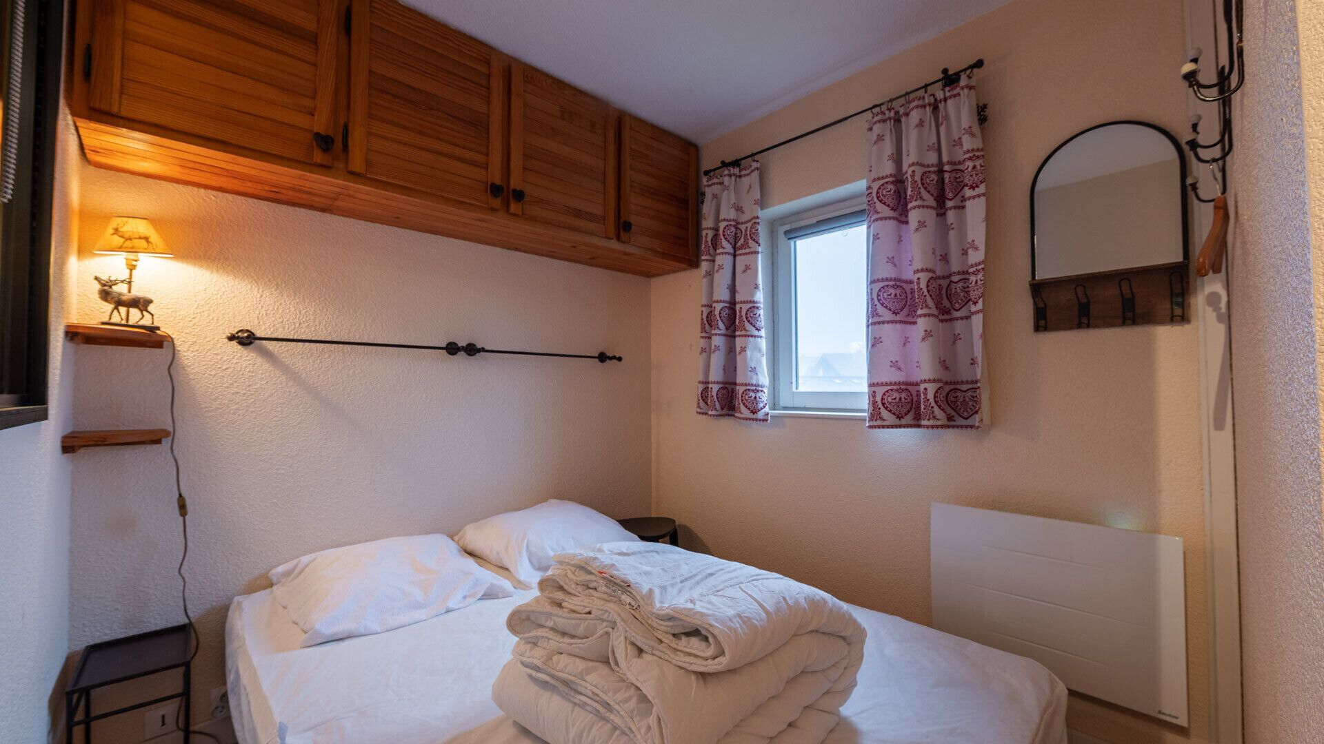 A bedroom with a bed, wooden cabinets, a window with patterned curtains, a wall-mounted lamp, and a radiator.