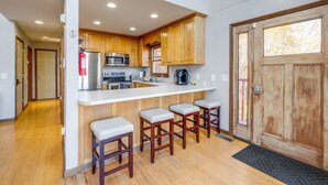 Una cocina moderna con armarios de madera, un refrigerador de acero inoxidable, un microondas y una cafetera. Hay tres taburetes de bar y un ventanal grande.