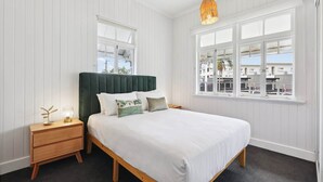 A bedroom with a bed, bedside tables, a hanging light fixture, and a window with a view of buildings. - Tranquil 3-Bed Family Home with Spacious Backyard (Nundah)