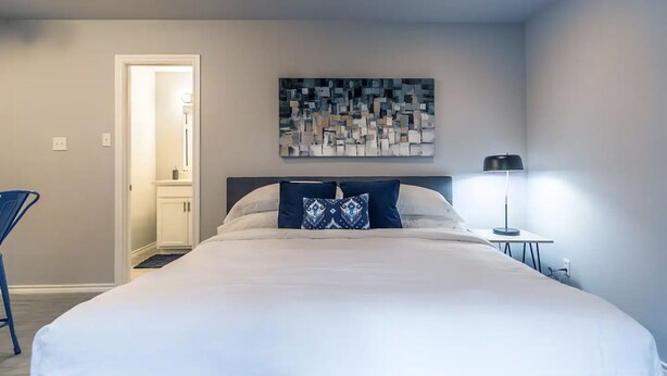 A bedroom with a large bed, a nightstand with a lamp, a door leading to another room, and a wall-mounted abstract art piece.