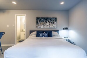 Une chambre avec un grand lit, une table de chevet avec une lampe, une porte donnant sur une autre pièce et une œuvre d’art abstraite accrochée au mur.