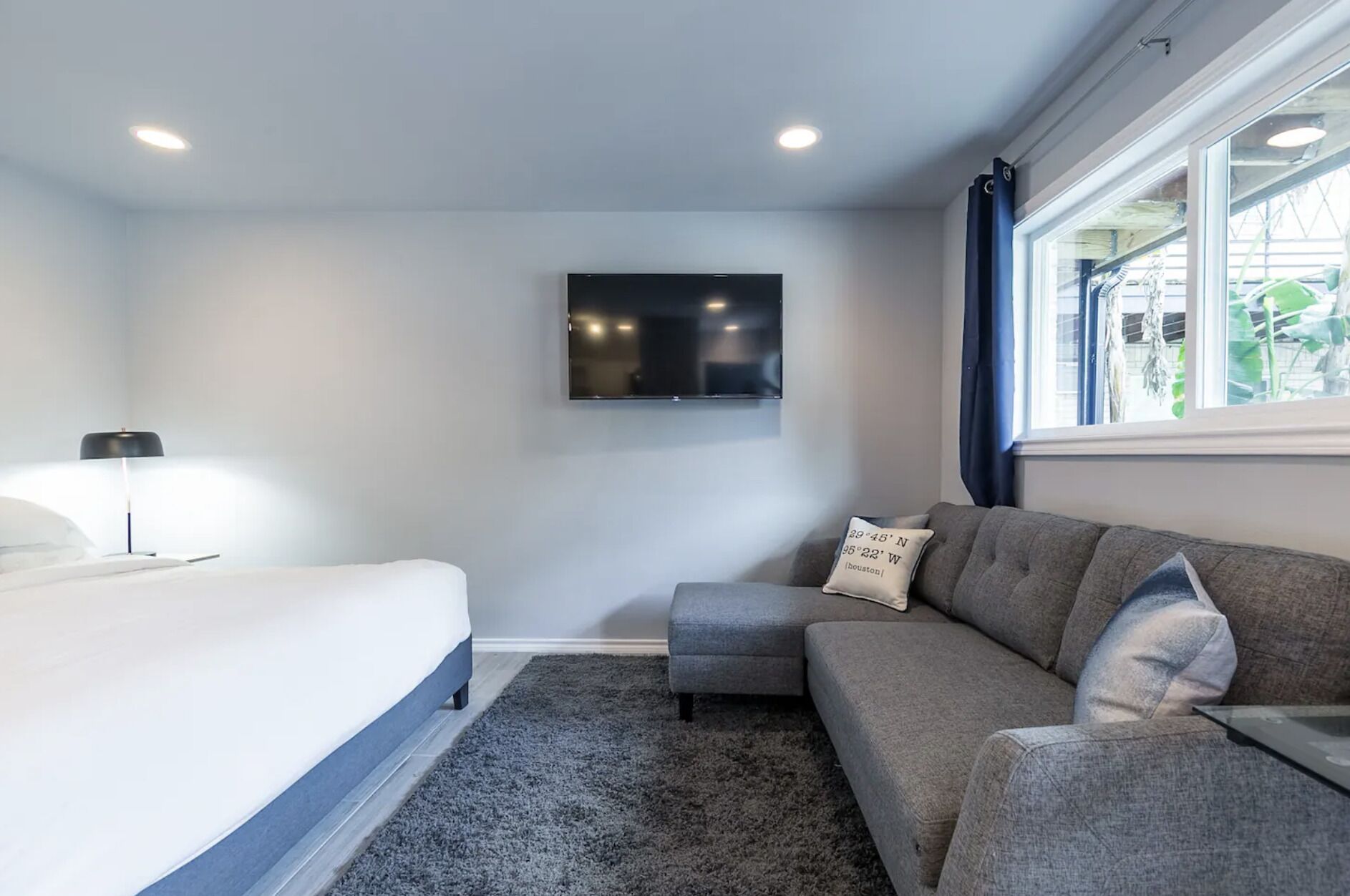 A modern hotel room with a bed, a grey sofa, a flat-screen TV mounted on the wall, and a window with curtains.